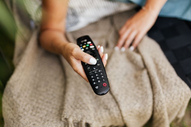 A person relaxing indoors, holding a TV remote control against a cozy blanket.