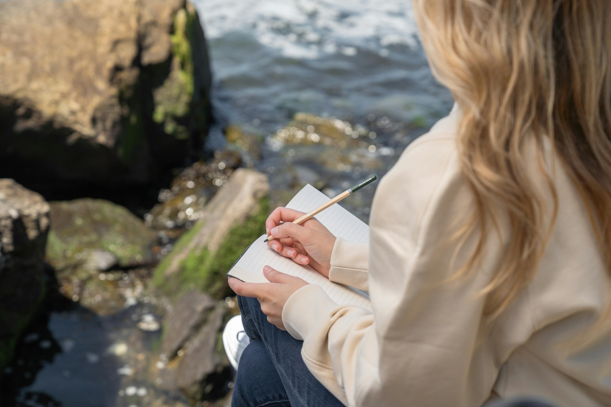 A woman sits by the rocky shore, journaling on a sunny day, reflecting the tranquility of nature.