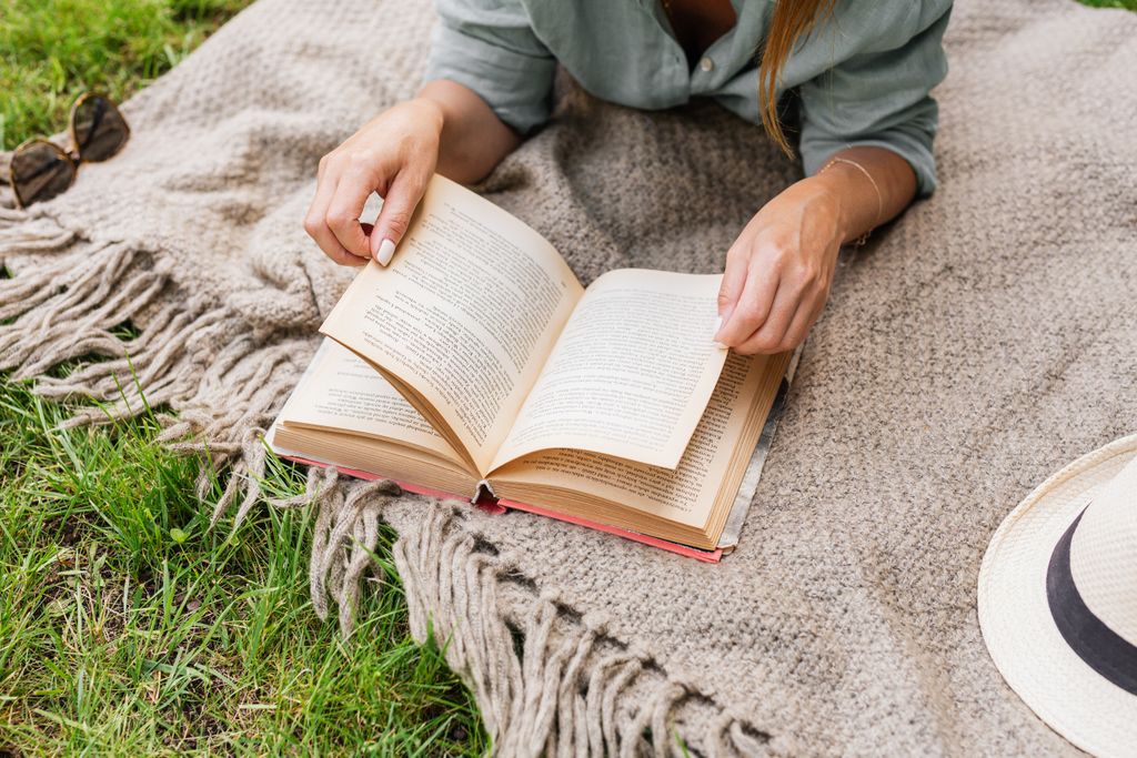 A woman enjoys a peaceful moment reading a book on a cozy blanket outside.